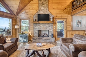 A cozy cabin interior with a stone fireplace, wooden walls, and a large window overlooking a scenic view.