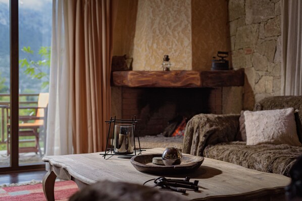 A cozy living room with a fireplace, a wooden table with a candle, and a view of greenery outside.