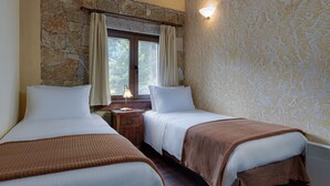 Une chambre avec deux lits, un mur en pierre, une fenêtre avec des rideaux et une table de chevet avec une lampe.