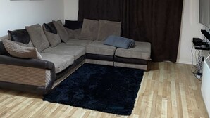 En stue med en hjørnesofa, et mørkt tæppe, trægulv, en loftlampe, et vægmonteret tv og et vindue med gardiner.