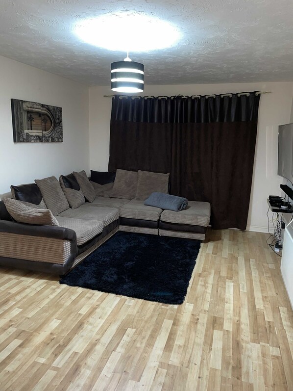 A living room with a sectional sofa, a dark rug, wooden flooring, a ceiling light fixture, a wall-mounted TV, and a window with curtains.