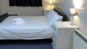 En pænt redt seng med hvide sengetæpper og et foldet håndklæde, et sengebord med en lampe, samt en vægmonteret lysarmatur.
