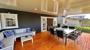 Un espacio al aire libre moderno con una terraza de madera, una mesa de comedor con sillas, un sofá con cojines y un portón corredizo que da a un porche cubierto.
