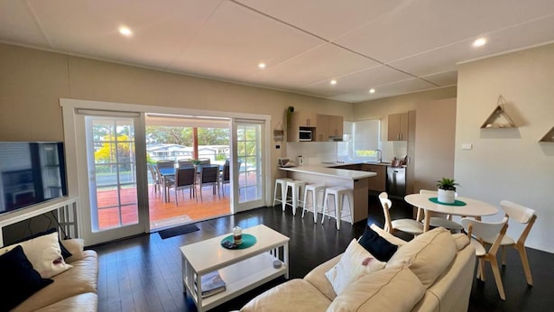 Una sala de estar moderna con un sofá, una mesa de centro y un televisor. Se ve una puerta corrediza de vidrio que da a un patio exterior con zona de comedor. Se observa la cocina, con armarios blancos y un microondas.