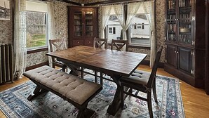 A dining room with a wooden table, cushioned benches, a ceiling fan, and a view of a house through large windows.