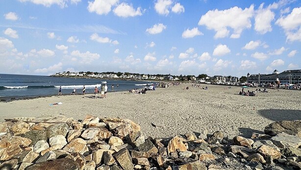 A beach with rocky foreground, a clear sky, and distant buildings.