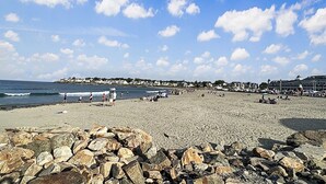 Una playa con primer plano rocoso, cielo despejado y edificios a lo lejos.
