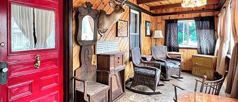 Interior em madeira com porta vermelha, cabeça de veado montada, móveis de vime e um candelabro.