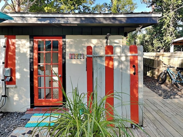 A small, single-story building with a red door and white walls, a wooden fence, and a bicycle parked outside.