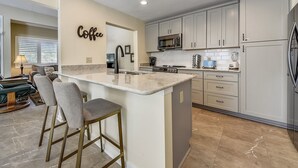 A modern kitchen with a central island, bar stools, a microwave, a refrigerator, and a coffee sign on the wall. - Spacious Pup Friendly Villa w/ Golf Course Views (Hilton Head Island)