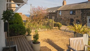 A courtyard with a wooden deck, potted plants, a wooden chair, and stone buildings in the background. - Craster Reach -  a family friendly that sleeps 6 guests  in 3 bedrooms (Alnwick)