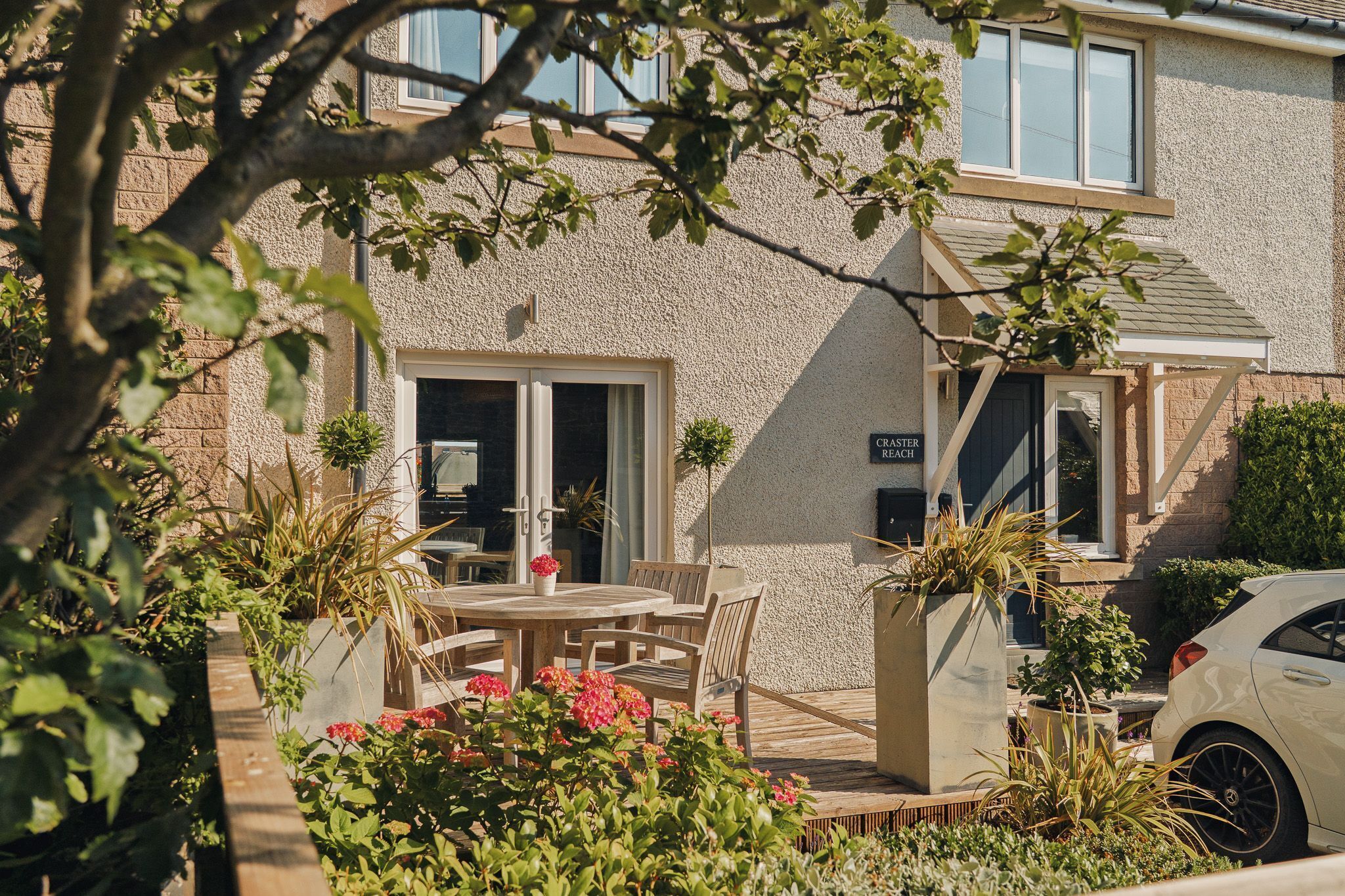 A residential building with a patio area featuring a wooden table and chairs, surrounded by potted plants and flowers.