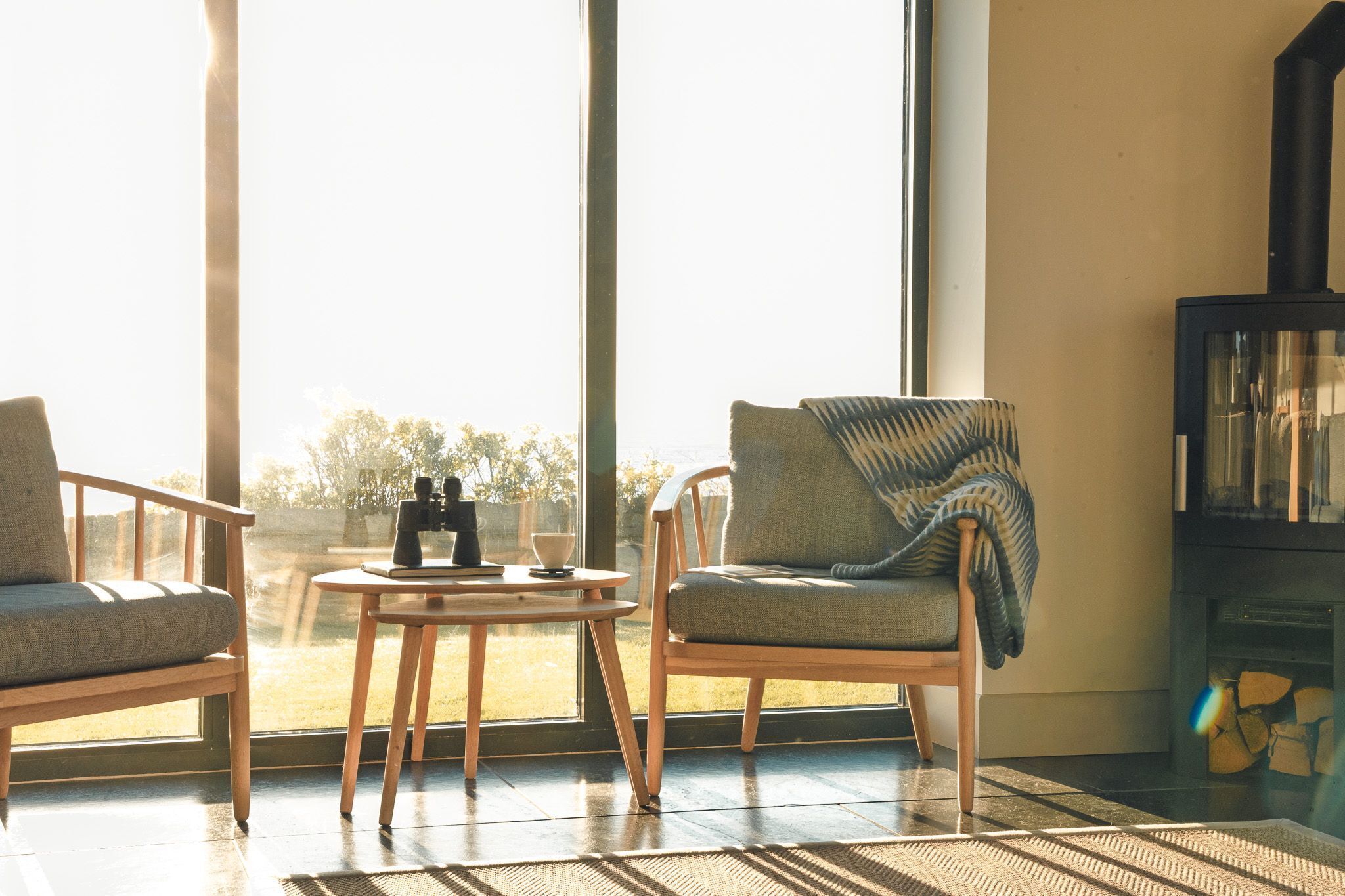 A room with two chairs, a small table, a fireplace, and a large window with a view of trees.