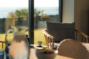 A room with a view of the sea, a chair, a table with a cup, and a window.