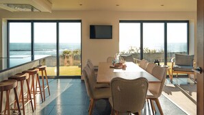 Un comedor con una mesa grande y sillas, un televisor montado en la pared y ventanas amplias con vista al océano.