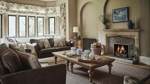 A cozy living room with a fireplace, a sofa, a coffee table, and a view of the outdoors through large windows. - Oughtershaw Hall -  a large propertie that sleeps 16 guests  in 8 bedrooms (Skipton)