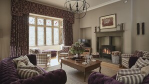 A living room with a fireplace, plaid furniture, and a large window with curtains. - Oughtershaw Hall -  a large propertie that sleeps 16 guests  in 8 bedrooms (Skipton)