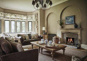 Un salon chaleureux avec une cheminée, un canapé, une table basse et une vue sur l’extérieur grâce à de grandes fenêtres.
