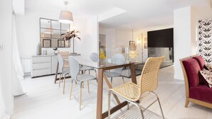 Ett modernt matområde med ett glasbord och stolar, en hängande lampa och utsikt in i ett annat rum.