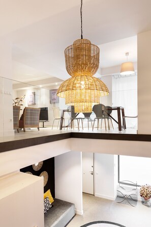 A modern interior with a dining area featuring a wicker pendant light, a sofa with cushions, and a view of a living room with a television and seating. - Genteel Home Francisco Silvela (Madrid)