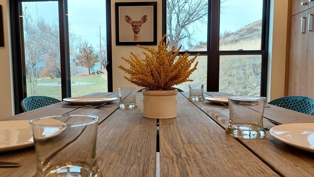 Une table à manger dressée avec des assiettes, des verres et une plante en pot, le tout agrémenté de grandes fenêtres et d’un tableau encadré au mur.