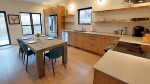 Une cuisine moderne avec des meubles en bois, un réfrigérateur en acier inoxydable, un lave-vaisselle et une cuisinière. On y trouve une table à manger avec des chaises et une fenêtre donnant sur les arbres.