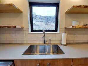 Una cucina con un lavello in acciaio inossidabile, una finestra sopra e scaffali di legno sopra il piano di lavoro.