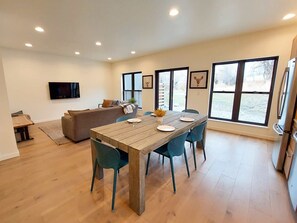 A modern living room with a wooden dining table, teal chairs, a sofa, and a TV mounted on the wall.