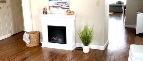 A living room with a fireplace, a painting of a sailboat, a potted plant, and a staircase leading to another room.