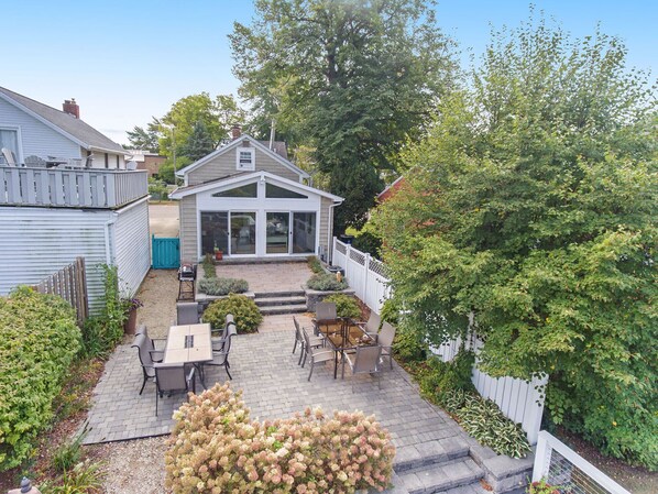 A house with a patio area featuring a table and chairs, surrounded by a white fence and lush greenery.