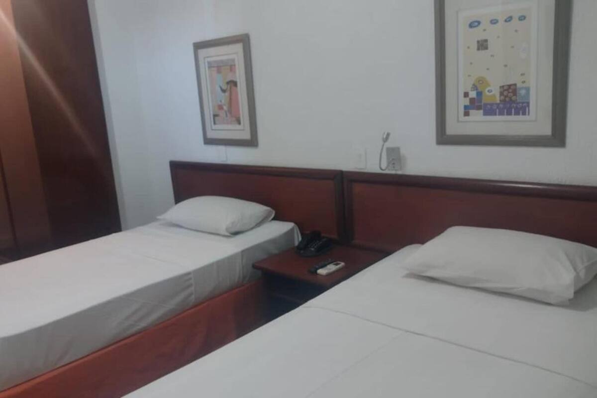 Quarto de hotel com duas camas de solteiro, cabeceiras de madeira, roupa de cama branca, mesa de cabeceira com telefone e tomadas na parede.