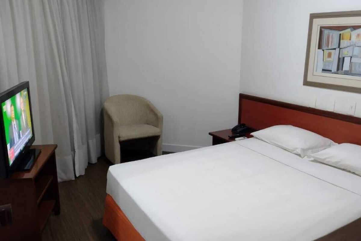 Quarto de hotel com cama, cadeira, televisão e um quadro na parede.
