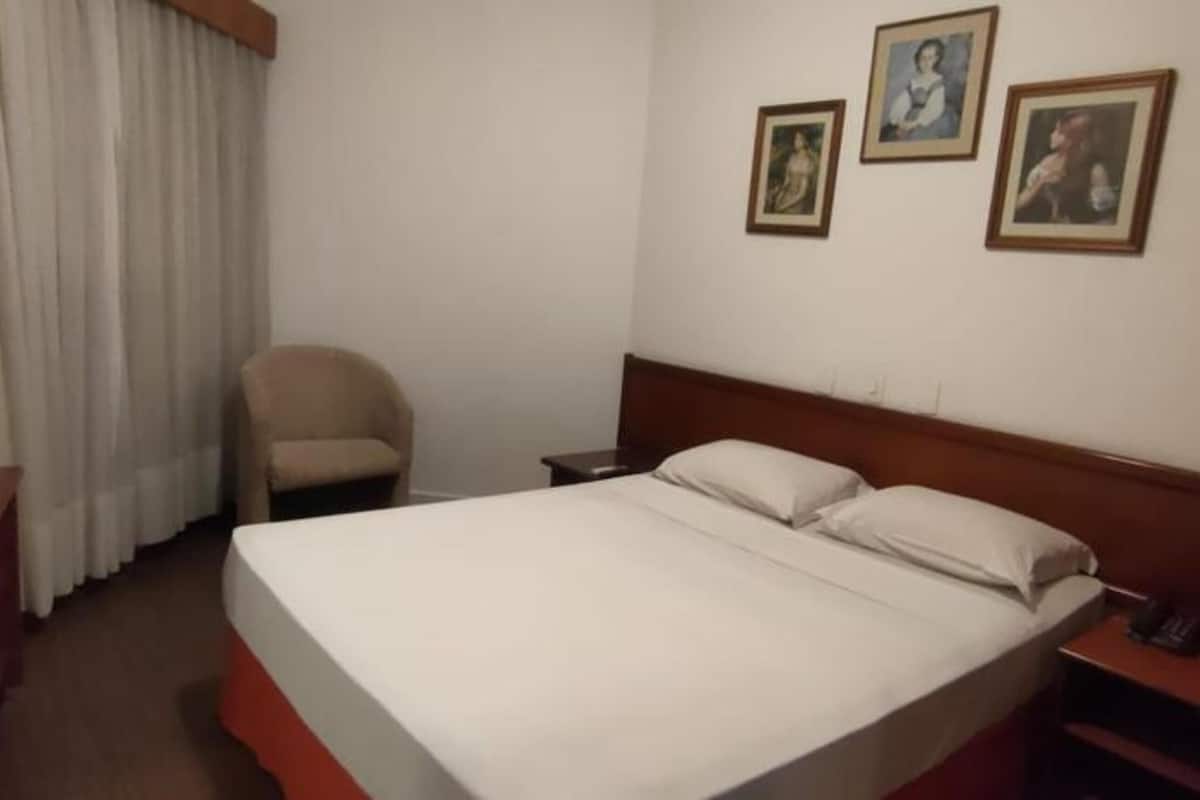 Quarto de hotel com cama, televisão sobre suporte, uma cadeira e três quadros na parede.