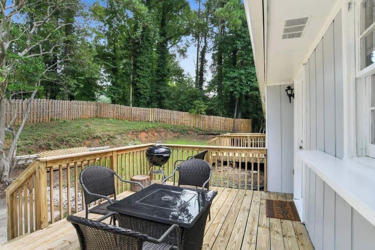 A wooden deck with a table and chairs, a barbecue grill, and a white building with a window.