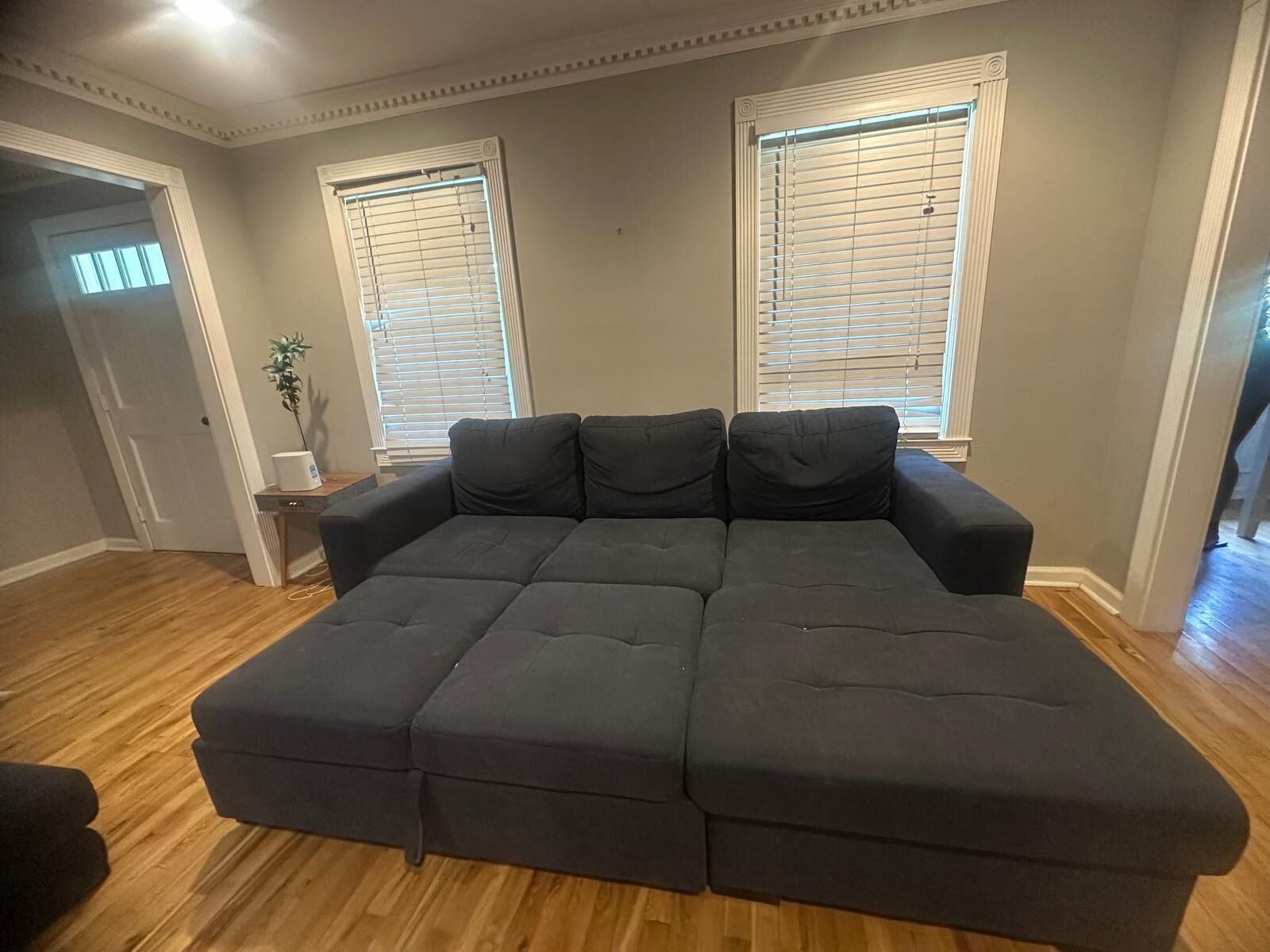 Una sala de estar con un sofá modular, dos ventanas con persianas, una mesita auxiliar con una planta y suelo de madera.