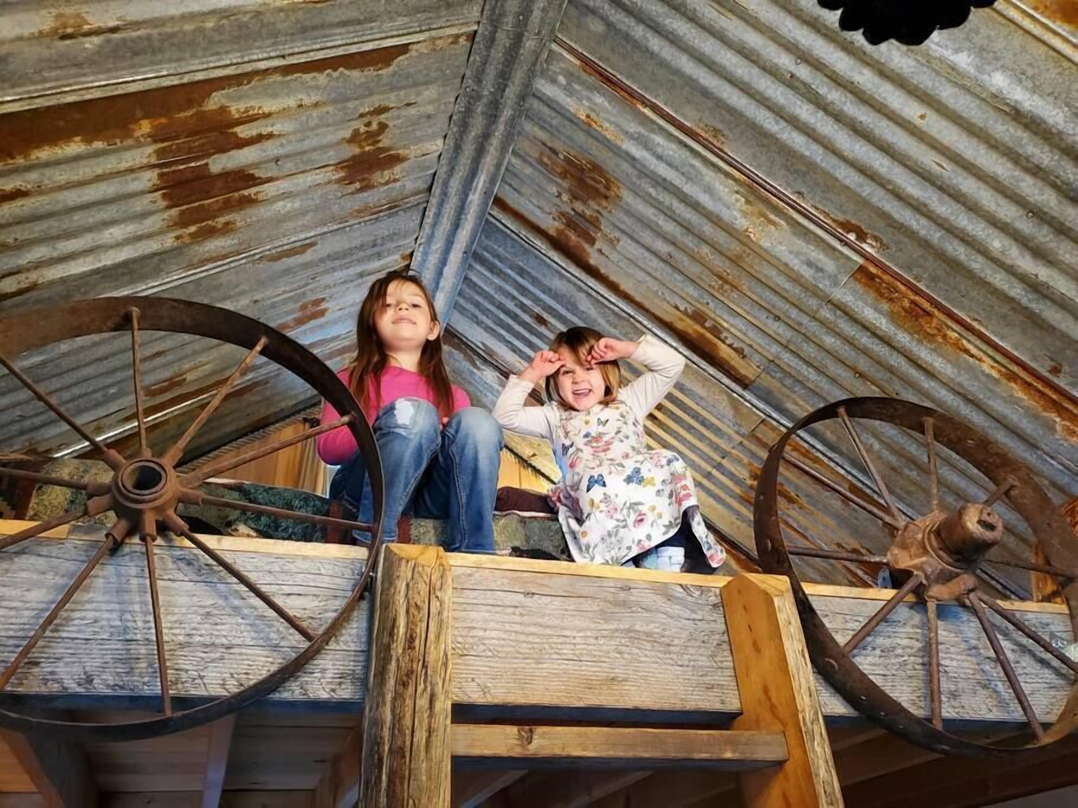 Twee kinderen zitten op een houten platform met grote, roestige wielen, onder een golfplaten dak.