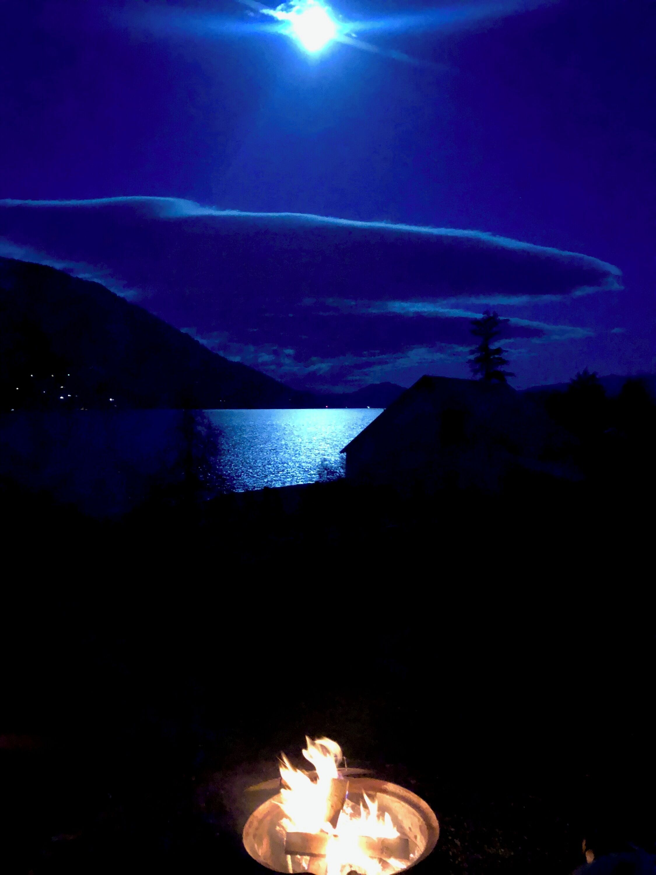 Een uitzicht over een meer in de nacht, met een vuurkuil op de voorgrond.