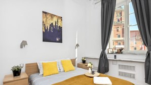 A modern hotel room with a bed, a nightstand, a window with curtains, a cityscape painting, and a view of buildings outside. - Przytulne apartamenty plac Zamkowy (Warsaw)