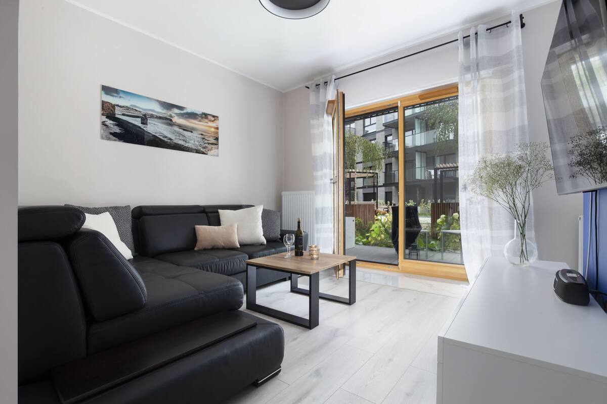Una sala de estar moderna con un sofá negro, una mesa de centro de madera y un ventanal con vistas a un edificio y zonas verdes.