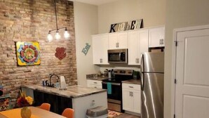 A modern kitchen with a brick accent wall, white cabinetry, stainless steel appliances, and a dining area with a wooden table and orange chairs. - Boutique Style Hotel -Entire Unit (Jeffersonville)