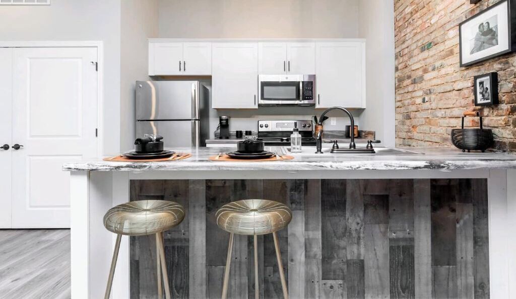 A modern kitchen with white cabinets, stainless steel appliances, and a marble countertop. There are two stools under the island.