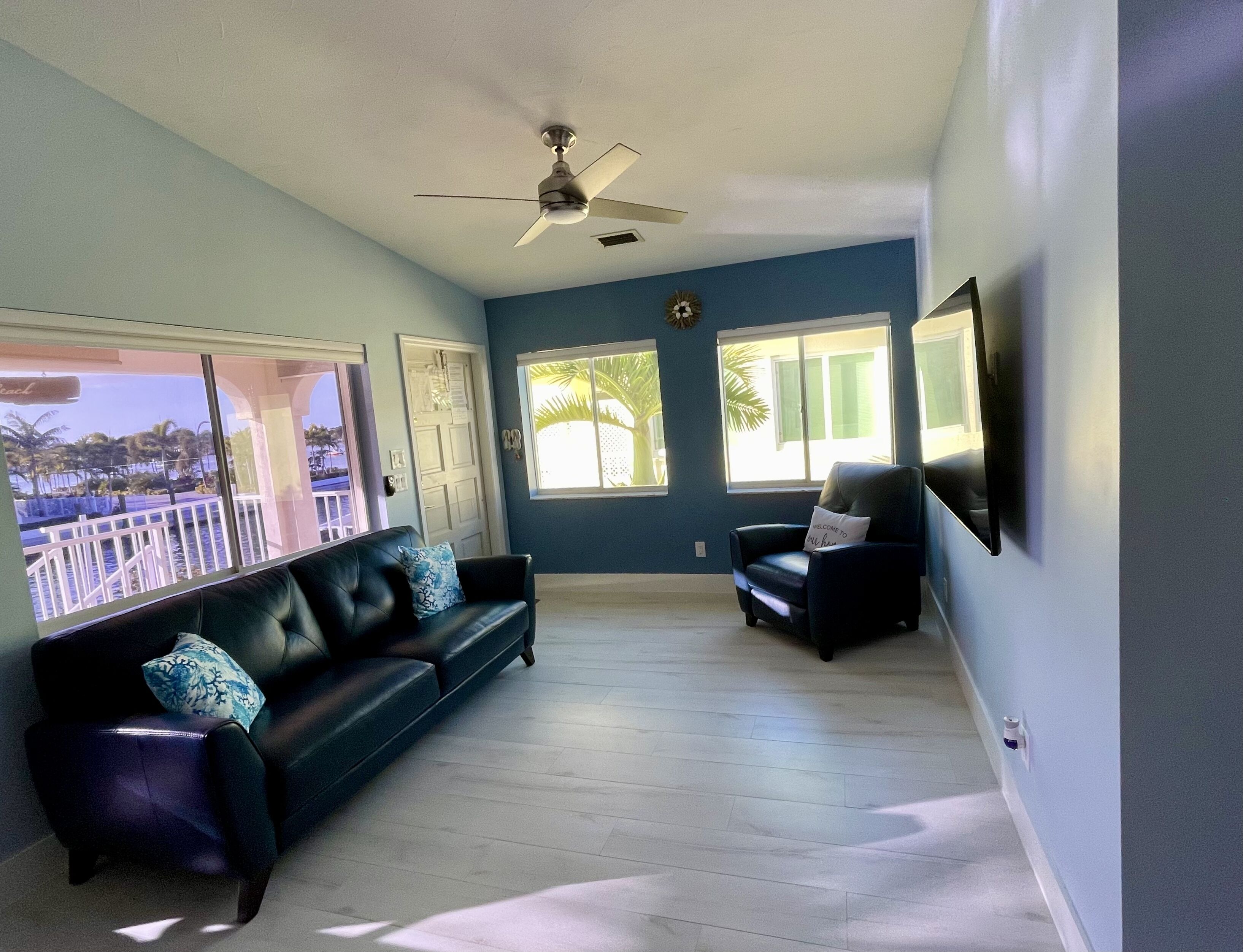 A spacious living room with a black leather sofa, a smaller armchair, and a ceiling fan. Large windows offer views of palm trees and a balcony.