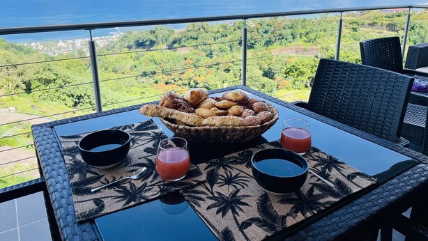 A table with a basket of pastries, two glasses of juice, and a bowl on a balcony overlooking a scenic view.