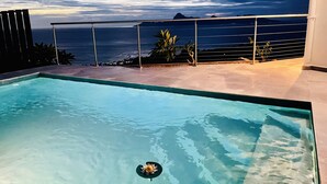 A swimming pool with clear water, a lotus flower, and a view of the ocean and mountains at dusk.