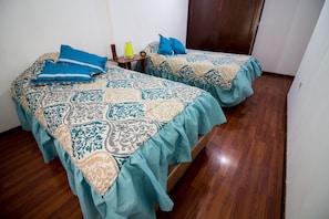 Two single beds with patterned bedspreads are positioned side by side, each accompanied by a nightstand with a lamp and decorative item.
