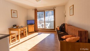 Una sala de estar acogedora con un sofá de madera, un televisor, una mesa de comedor y un amplio ventanal corredero que da a un espacio exterior.