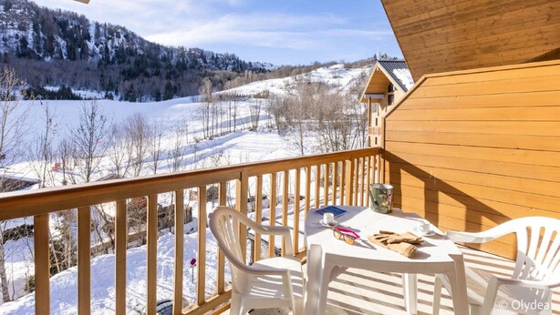 Una terraza de madera con una mesa y sillas, con vistas a un paisaje nevado con árboles y un edificio al fondo.