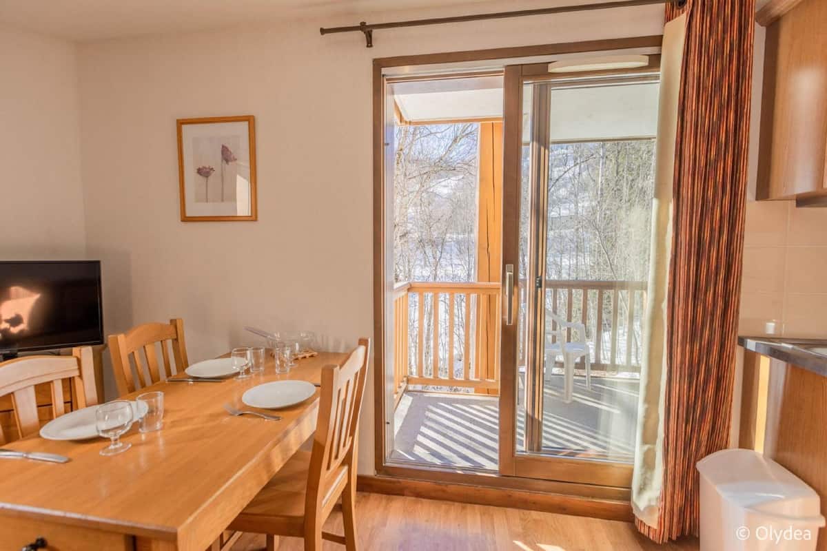 Un comedor con una mesa de madera preparada para cuatro personas, un televisor a la izquierda y un ventanal corredizo que da a un balcón exterior.