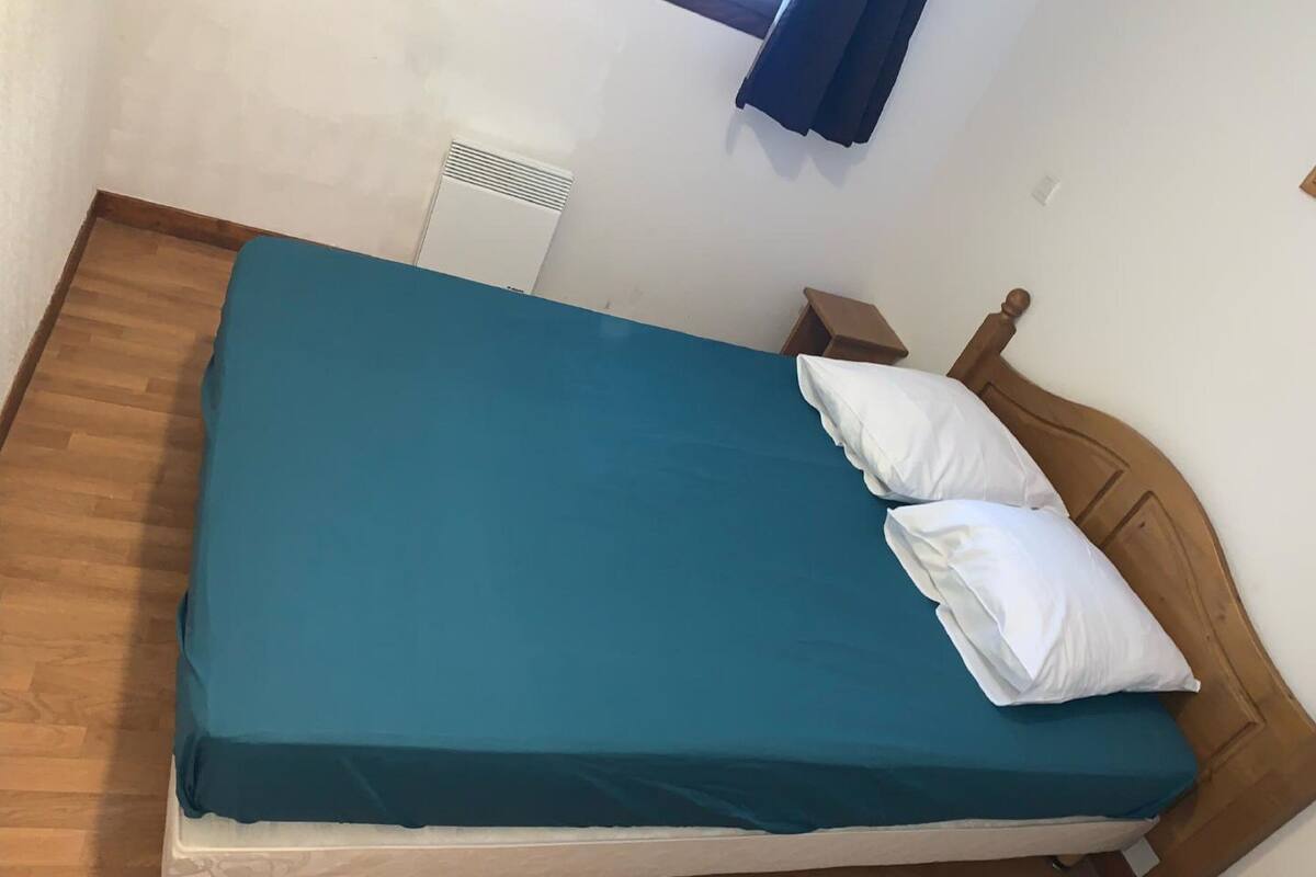 Una cama individual con una colcha color turquesa y dos almohadas blancas en una habitación con piso de madera y pared blanca.