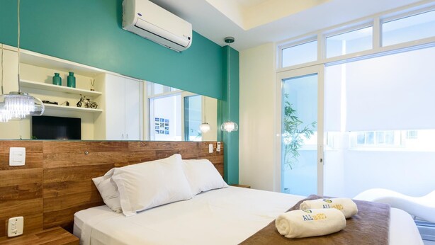 A hotel room with a wooden headboard, a large bed with white linens, a brown throw blanket, and a large window with a view of the outside. - Side sea View 3min From the Beach Pj204 (Rio de Janeiro)